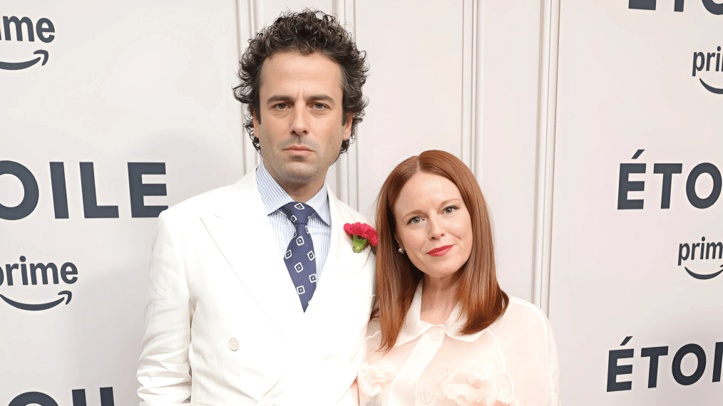 Luke and Andrea posing on a red carpet, with one in a white suit and the other in a light-colored dress, at a premiere event backdrop.