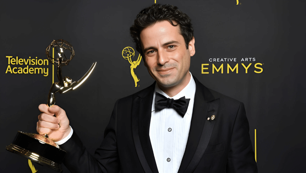 Luke Kirby in a black tuxedo holding an Emmy award while smiling in front of the Creative Arts Emmys backdrop.