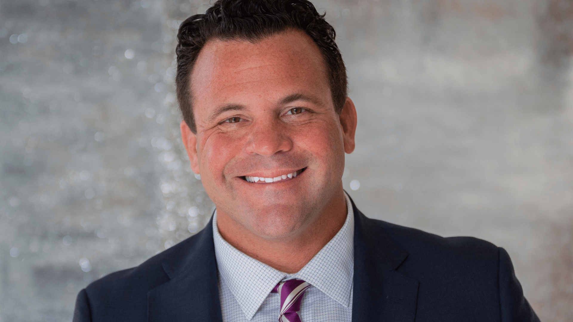 John Cerasani in a navy suit, light checkered shirt, and striped purple tie against a softly blurred gray background.