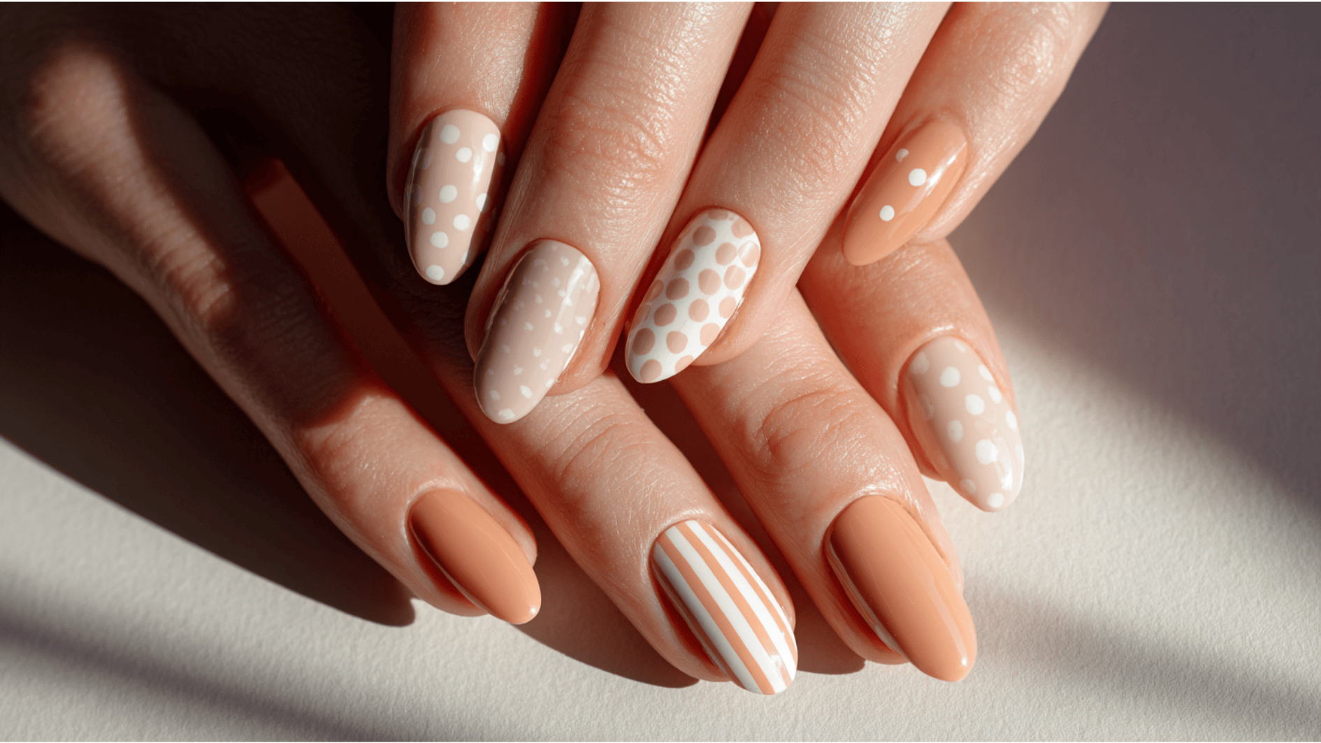 A woman's hands display an asymmetrical nail design featuring orange and white patterns, including stripes and solid colors