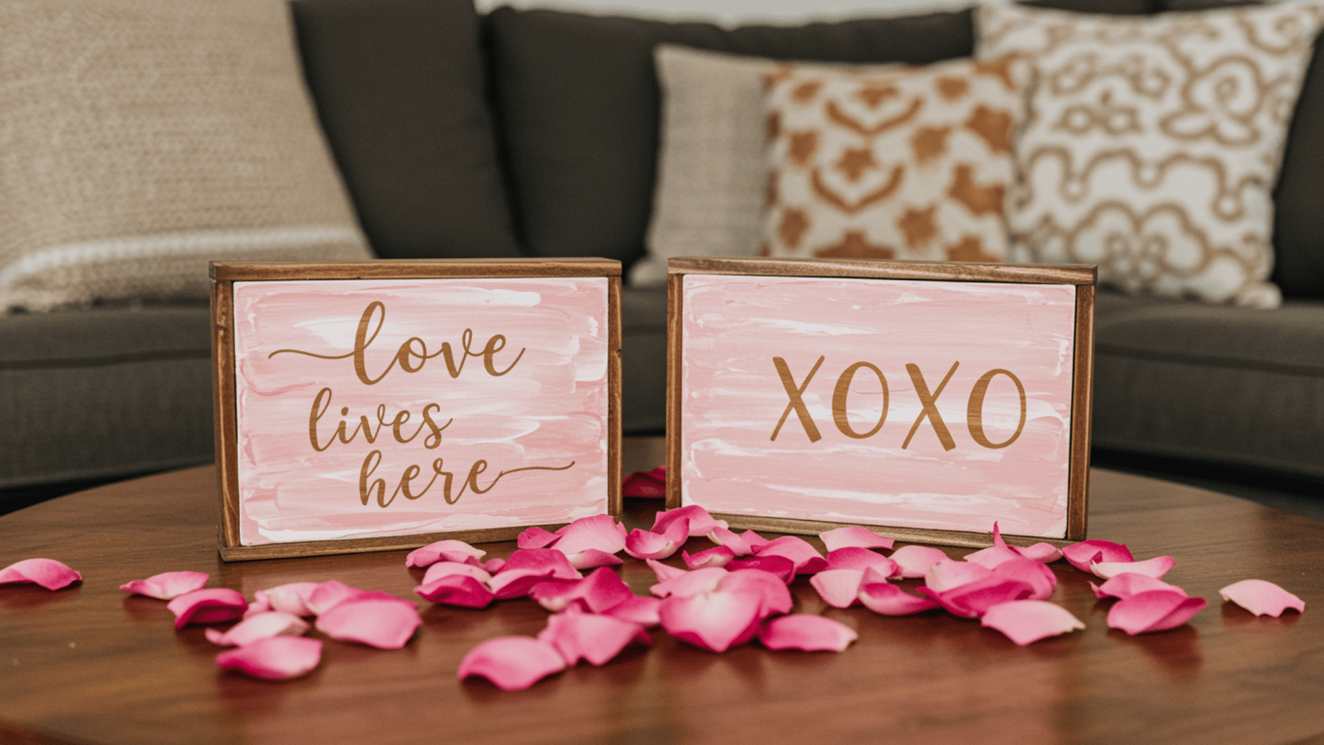 Two wooden signs with pink and white painted backgrounds reading love lives here and XOXO rest on a dark wood table scattered with bright pink rose petals.