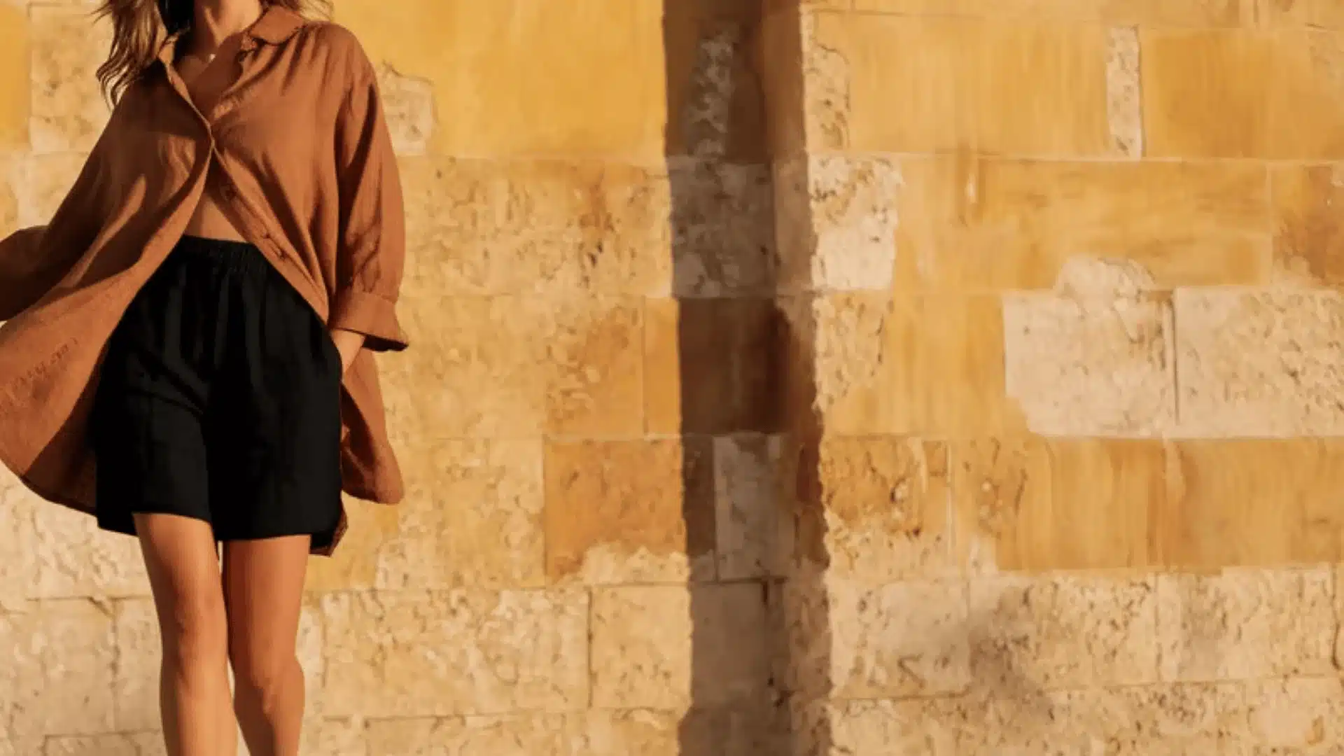 Woman wearing a flowing brown linen shirt open over black shorts stands against a textured sunlit yellow stone wall.