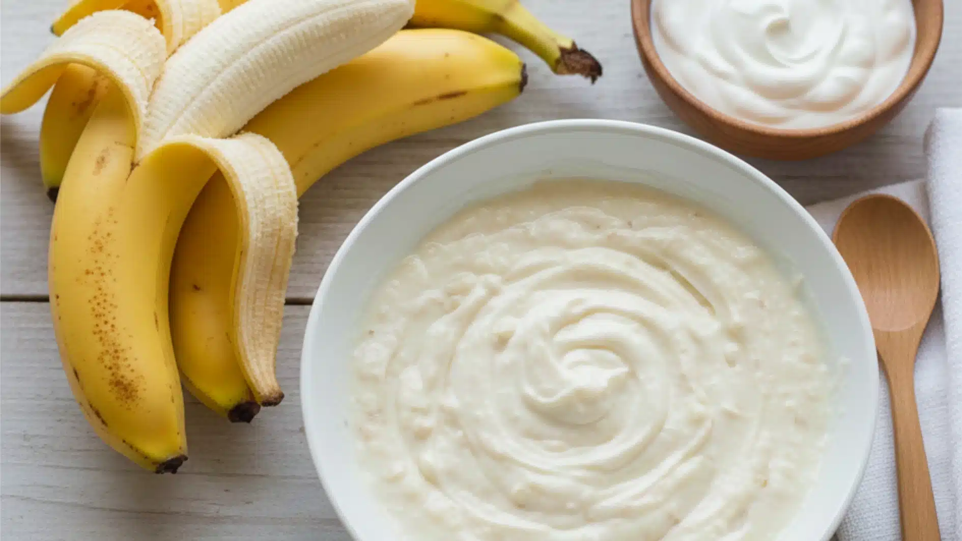 A bowl of creamy banana puree sits on a light wooden surface next to peeled bananas and a small bowl of yogurt