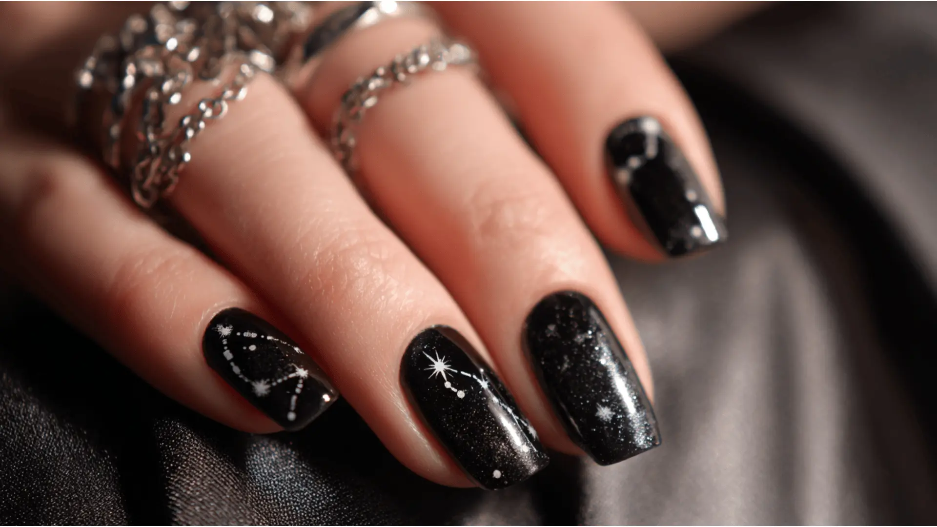 A close-up of a woman’s hand with glossy black nails featuring star constellation patterns creating a cosmic night sky inspired nail design