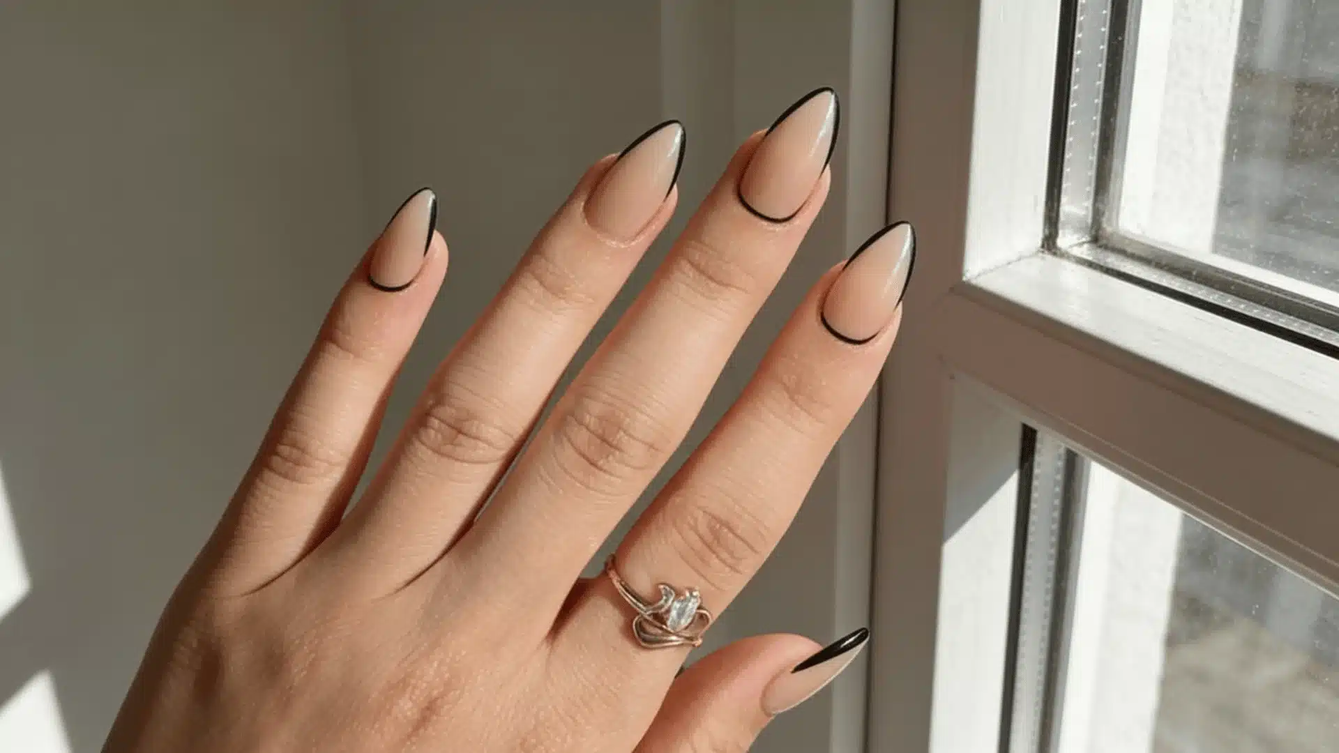 A close-up of a woman's hand with nude nails with micro black outline complemented by a decorative ring showcasing stylish manicure