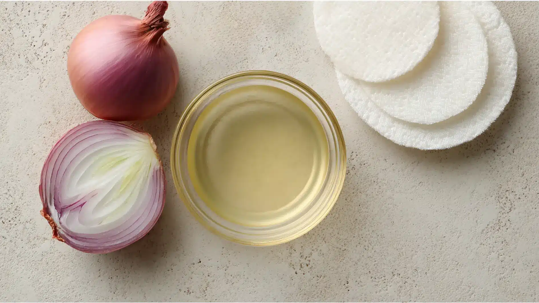 A whole red and halved red onion a small bowl of pale yellow oily and a stack of white cotton pads are arranged on a light textured surface.