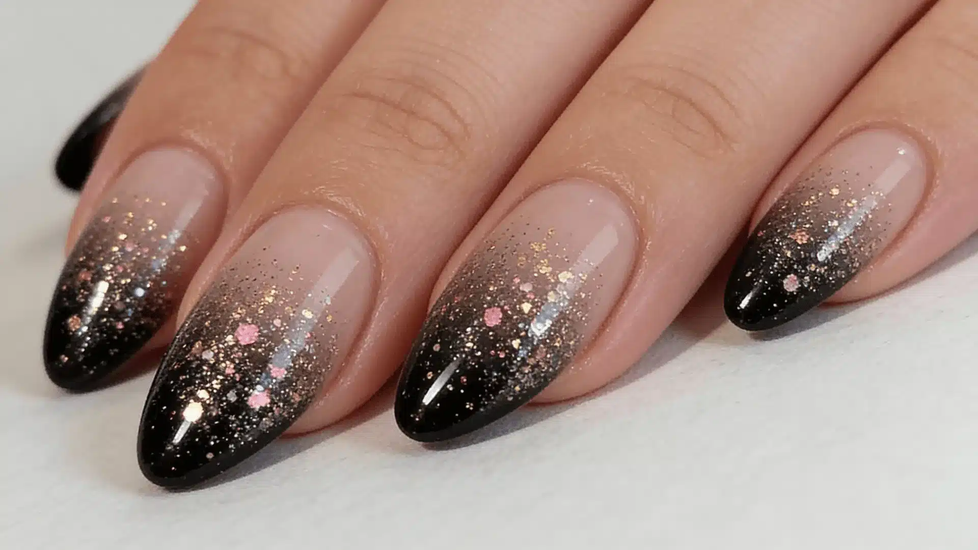 A woman's hand displaying nails with a nude and black glitter ombre effect and gold accents with a natural white background