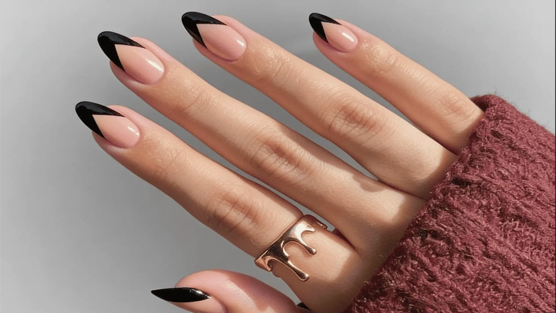 A woman's hand displaying nude nails with stylish black chevron tips with a stylish gold ring with a plain white wall as background