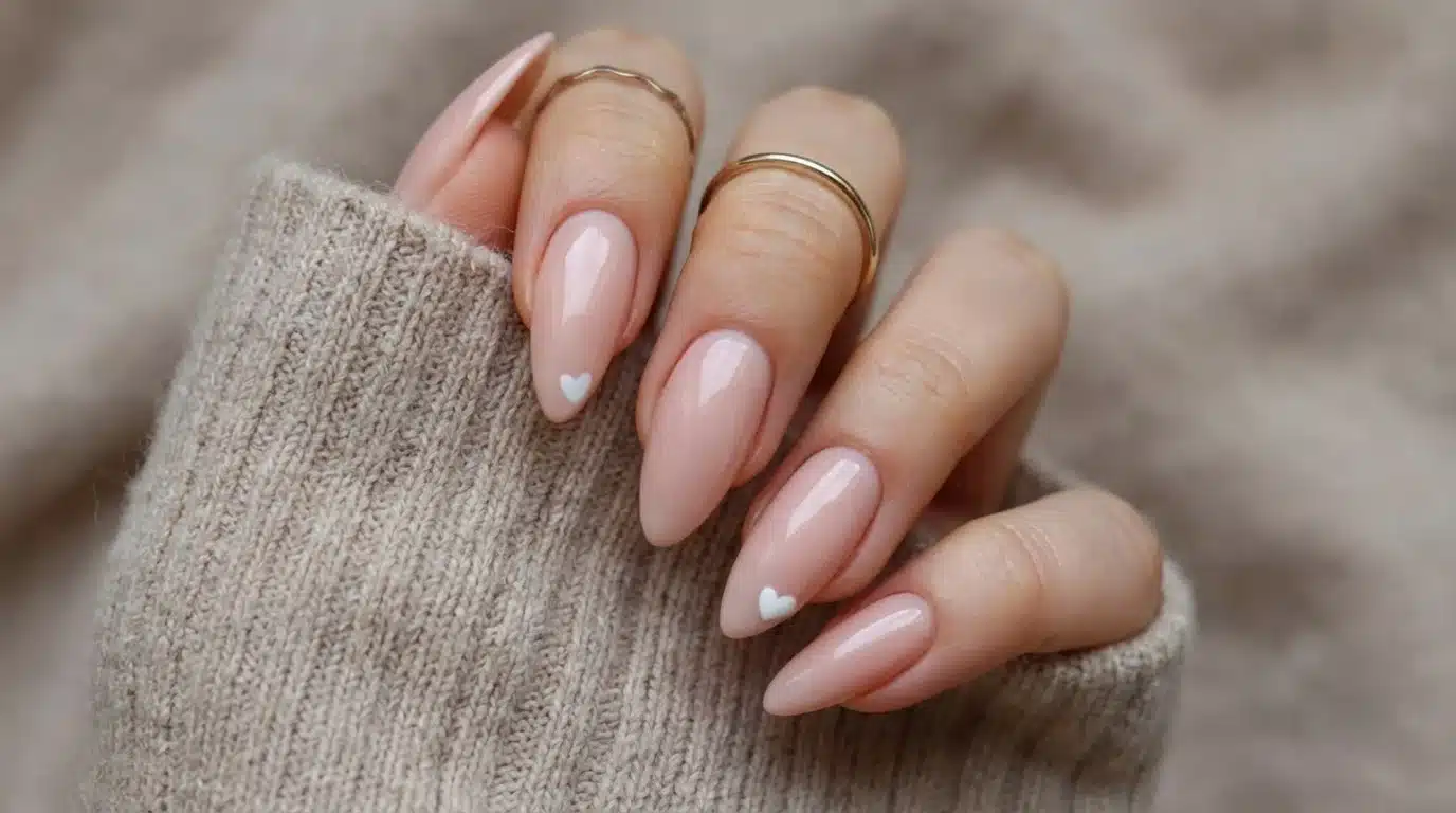 A woman's hand with nude nails accented by a heart design and a ring, highlighting a chic and polished look