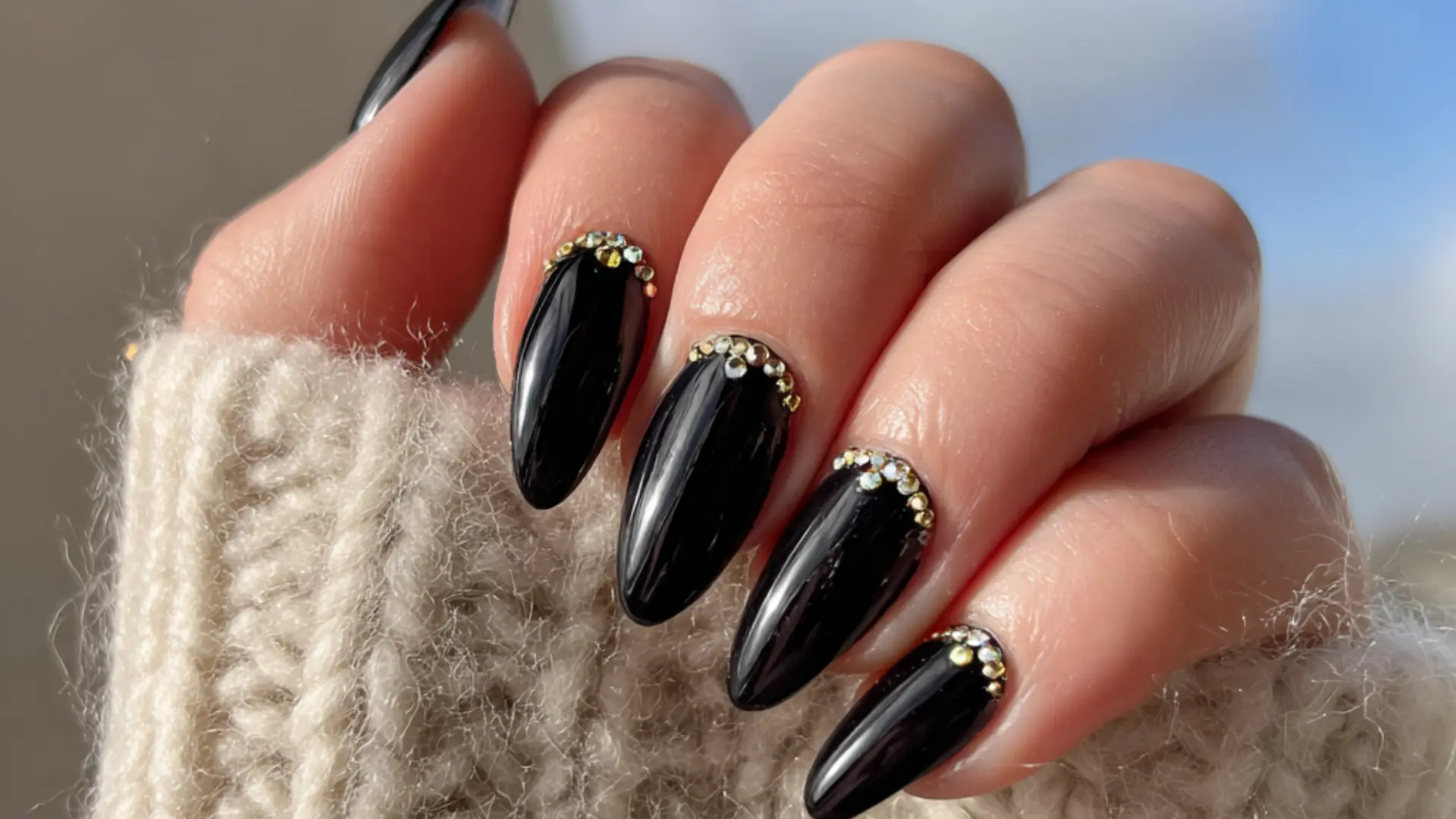 Black stiletto nails featuring gold accents and tiny gems at the cuticles, perfect for a chic manicure paired with glossy nail paint