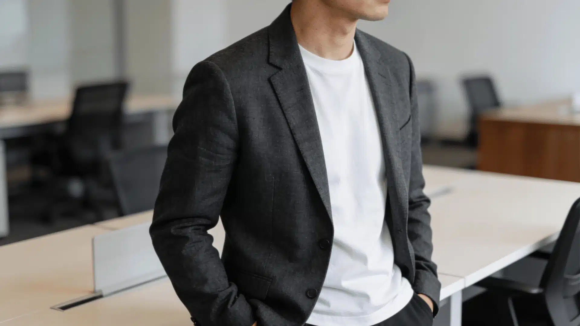 Close-up of a casually dressed professional man wearing a charcoal blazer over a white t-shirt standing in a modern, softly lit office environment.