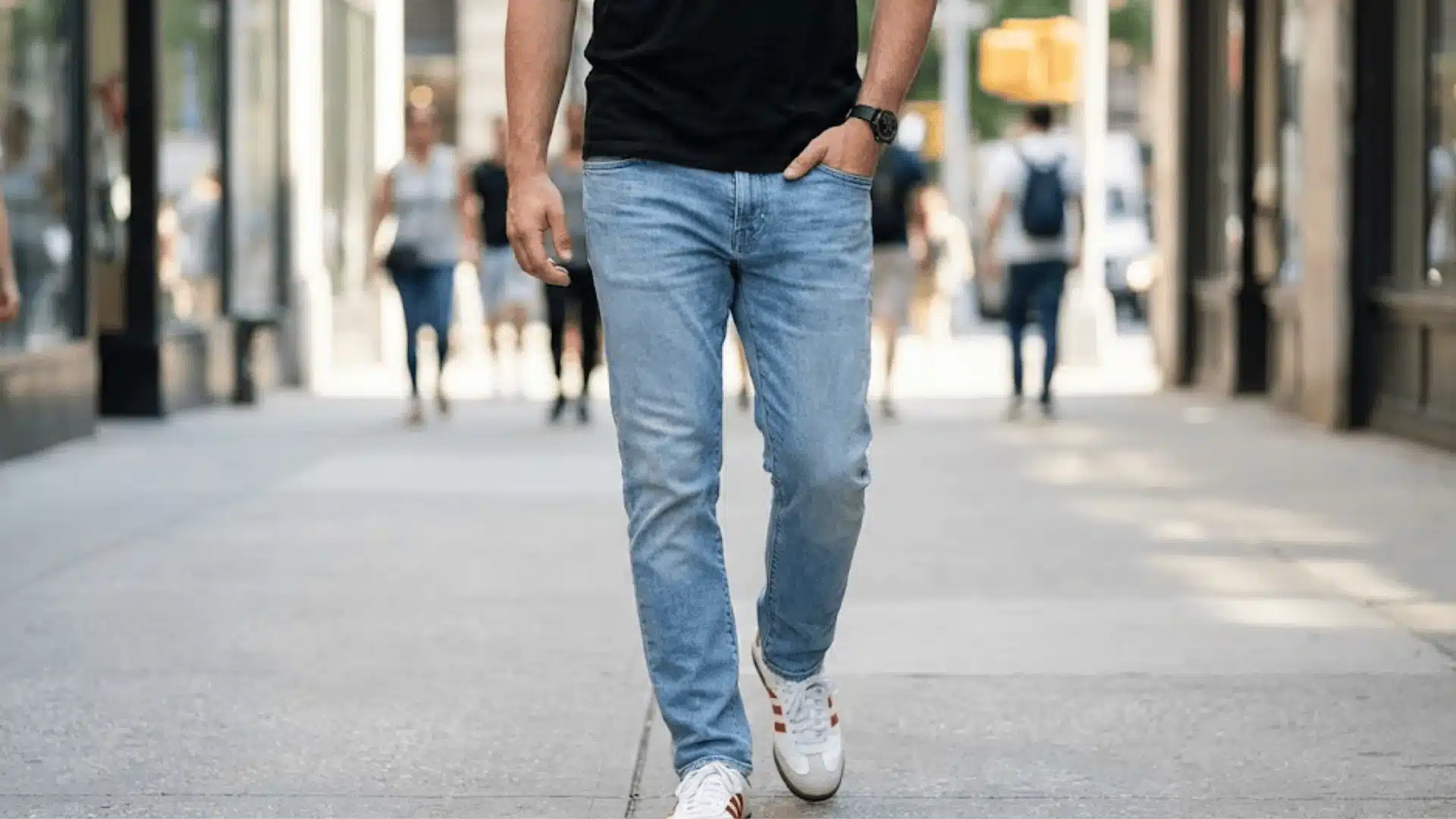 Man wearing light wash jeans, a black t-shirt, and white sneakers walking on a city sidewalk with blurred pedestrians in the background.