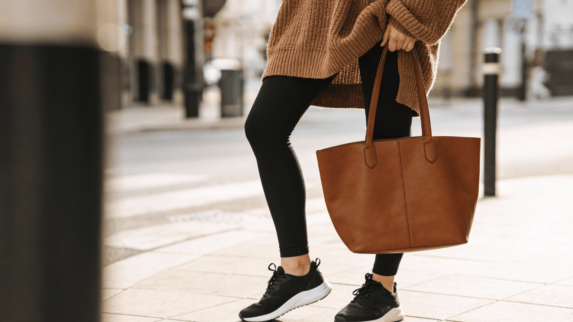 A woman wearing a brown knit sweater, black leggings, and black sneakers, holding a brown leather tote bag on a city sidewalk.