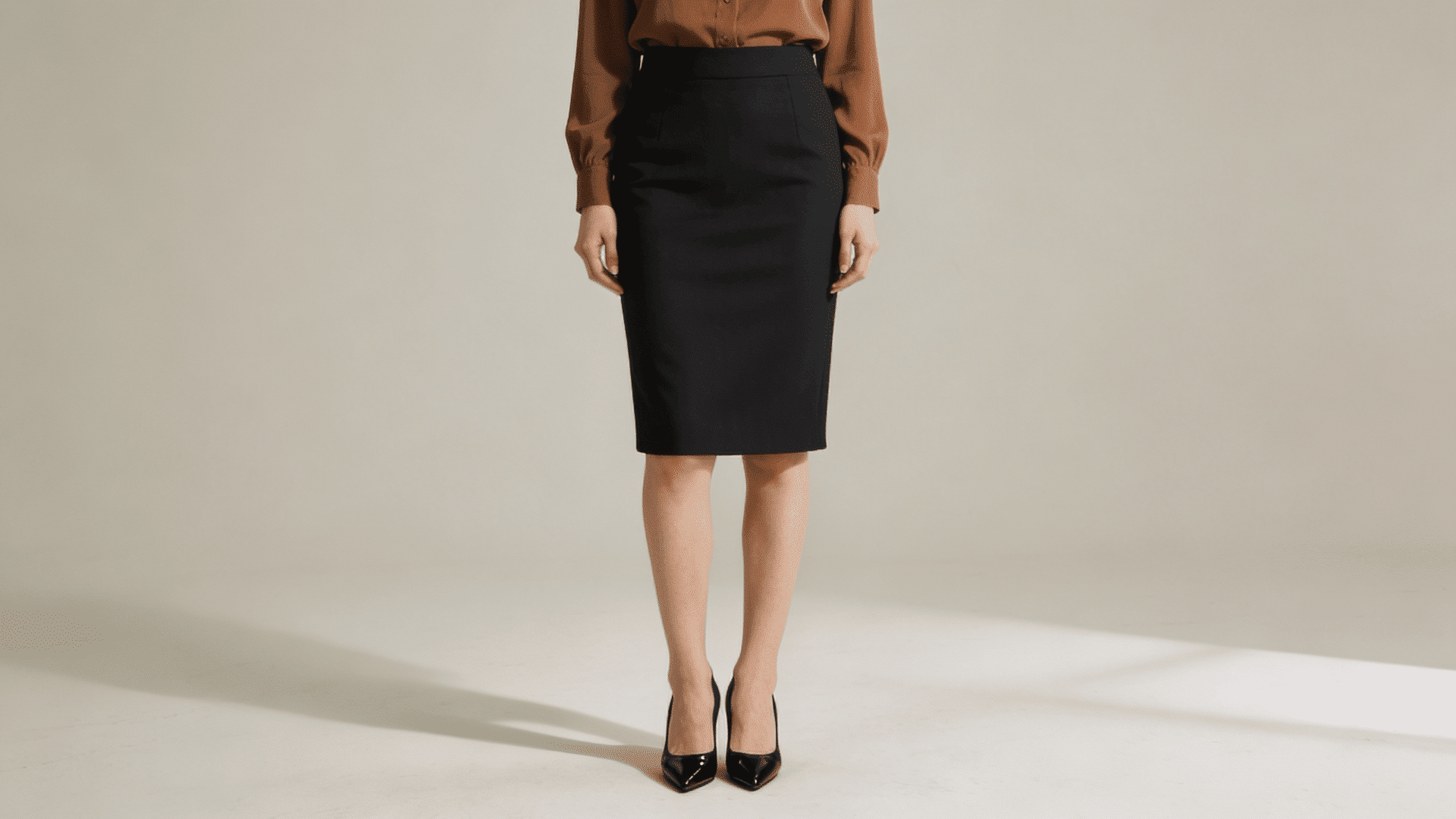 A woman wearing a fitted black pencil skirt, a brown blouse, and black patent leather heels against a light gray background.