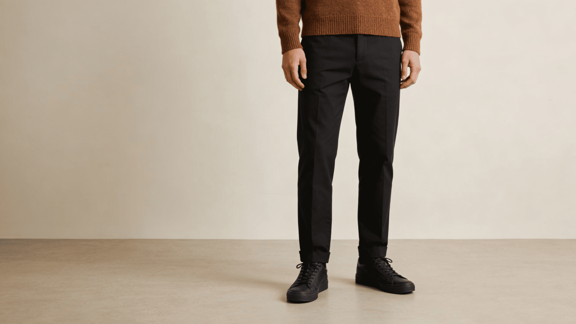 Man wearing a brown sweater, black trousers, and black lace-up sneakers against a neutral beige background.