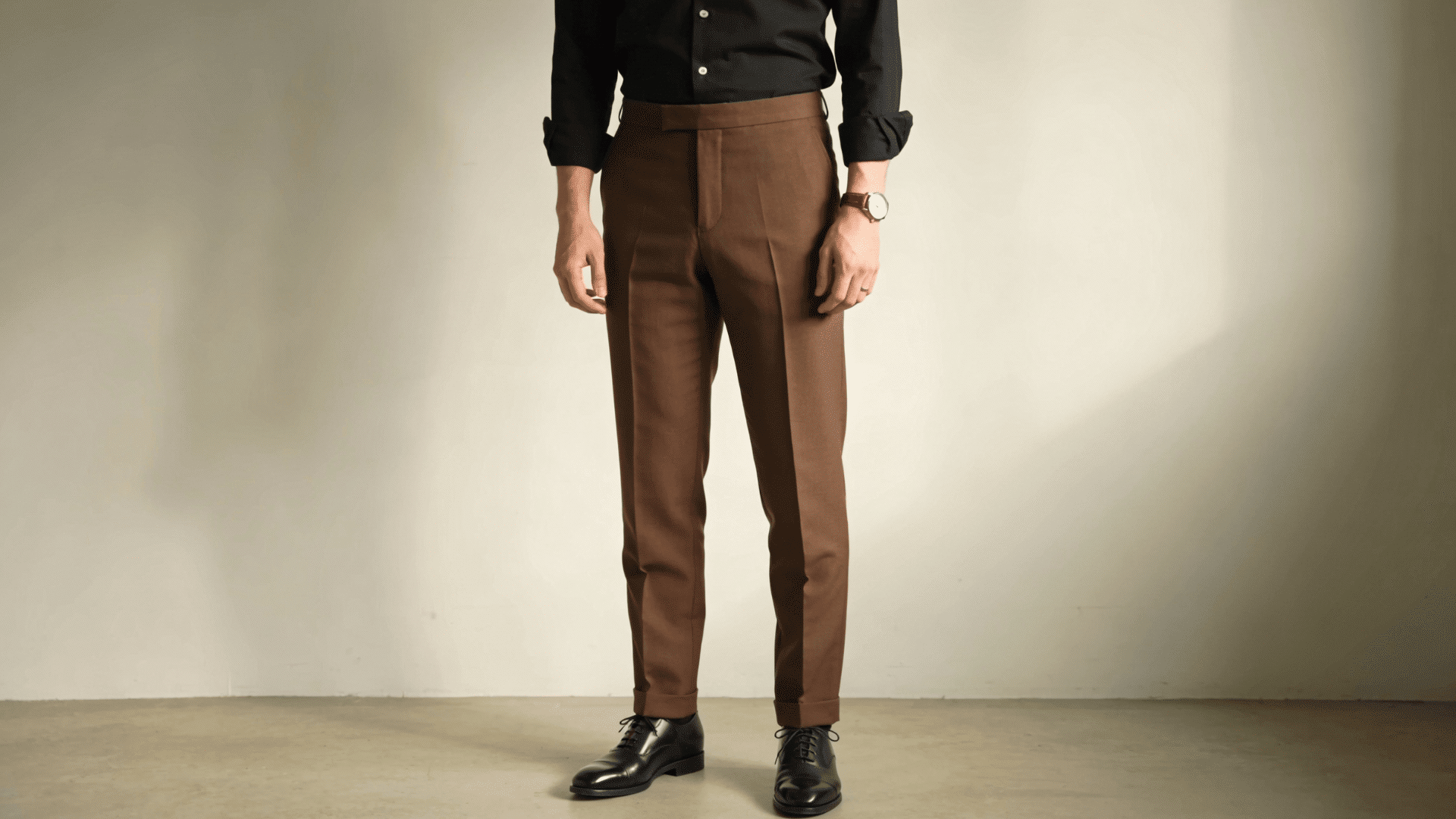 Man wearing high-waisted brown trousers, a black button-down shirt, and black polished leather Oxford shoes against a beige wall.