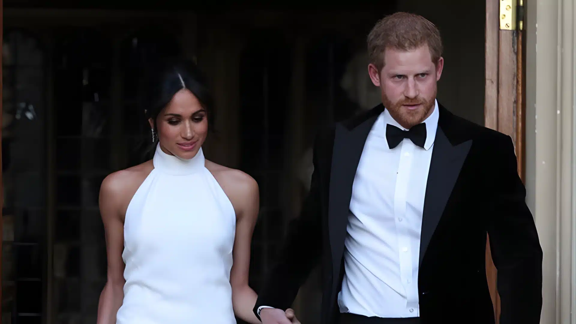 Meghan Markle in a white halter gown holds hands with Prince Harry in a black tuxedo, exiting a doorway.