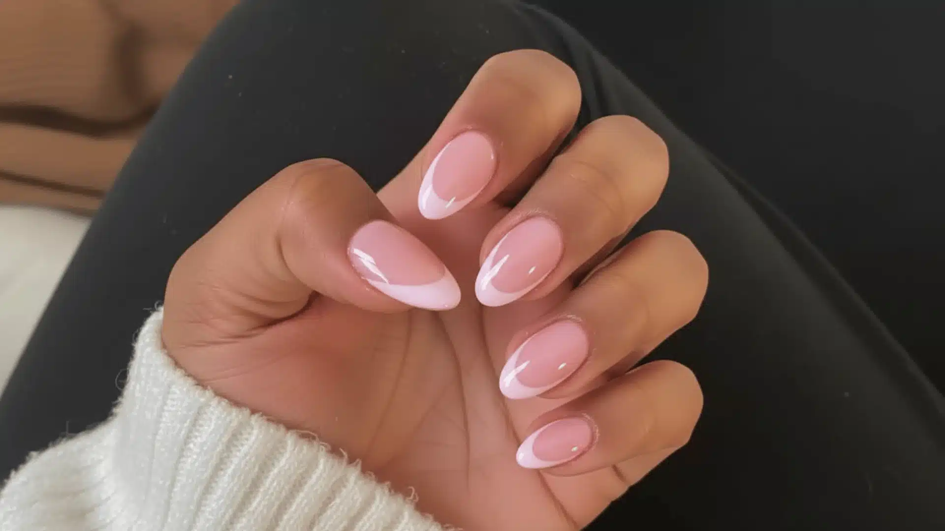 A woman with French tip on pink white nails in almond shaped holds a wearing white sweater showcasing her chic and stylish manicure