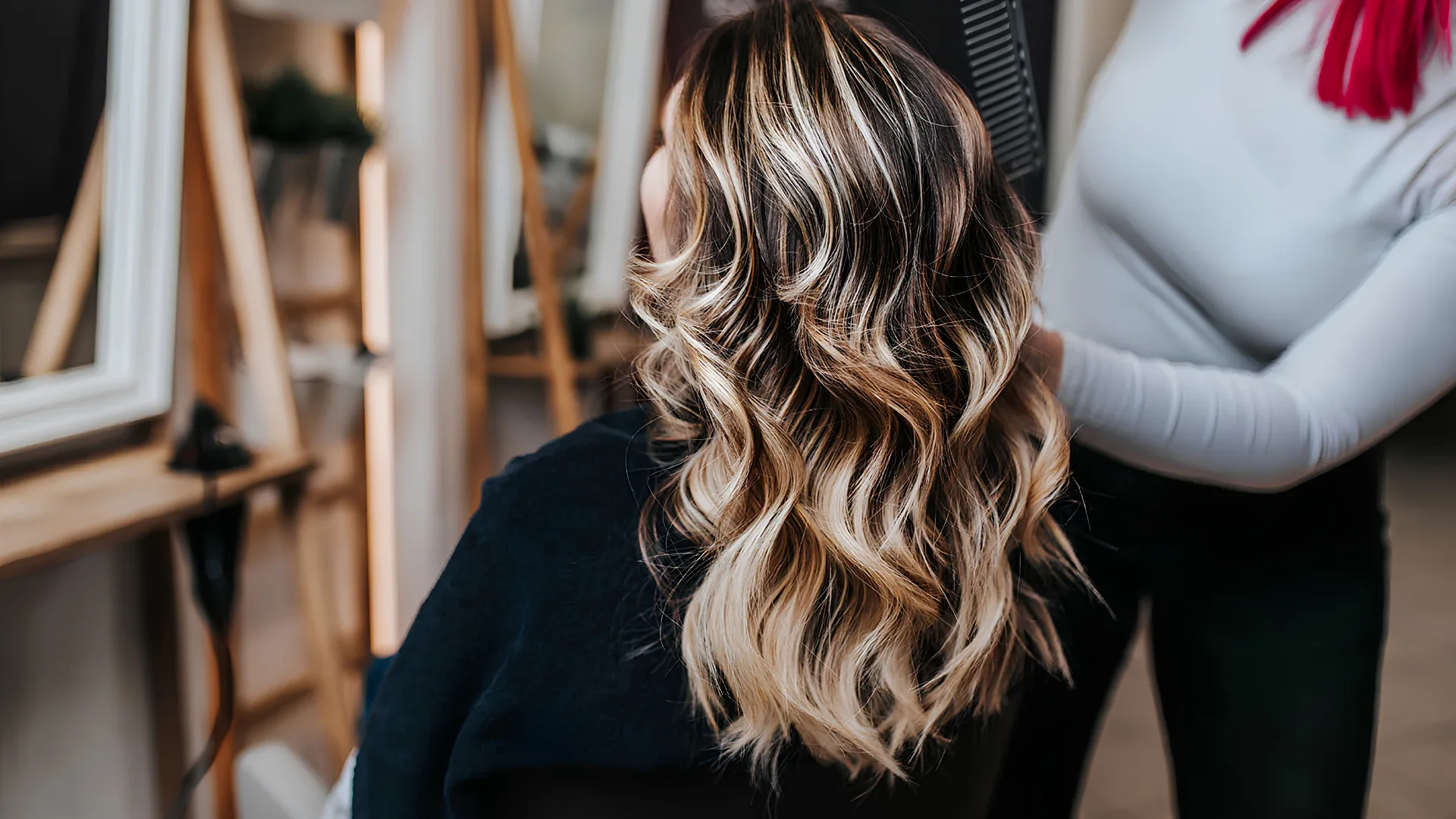 A person with long, wavy blonde and brown balayage hair, with a hairstylist's hands in the background