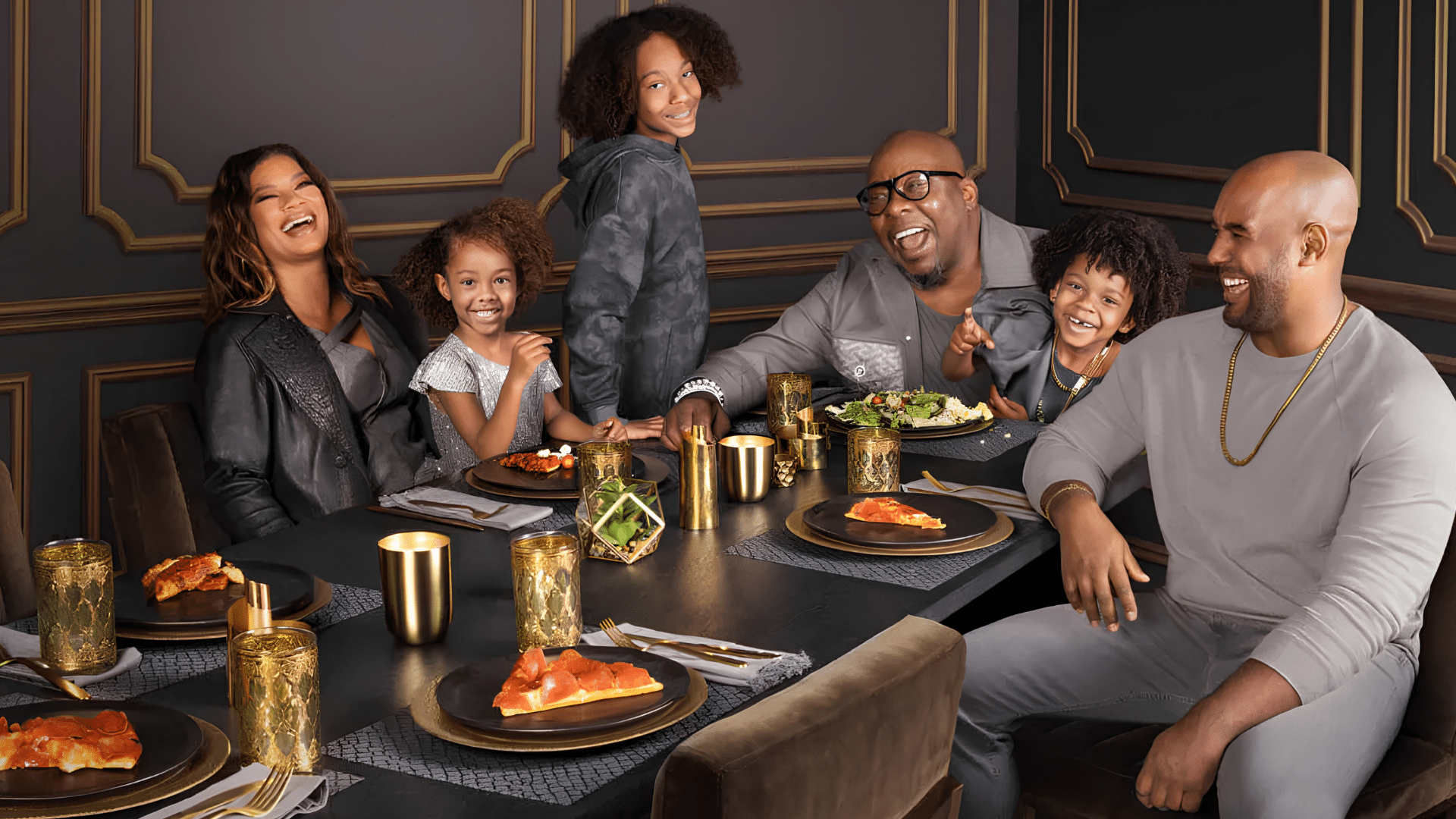 Bobby Brown with his family of six is laughing around a dinner table set with pizza, salad, and gold accents