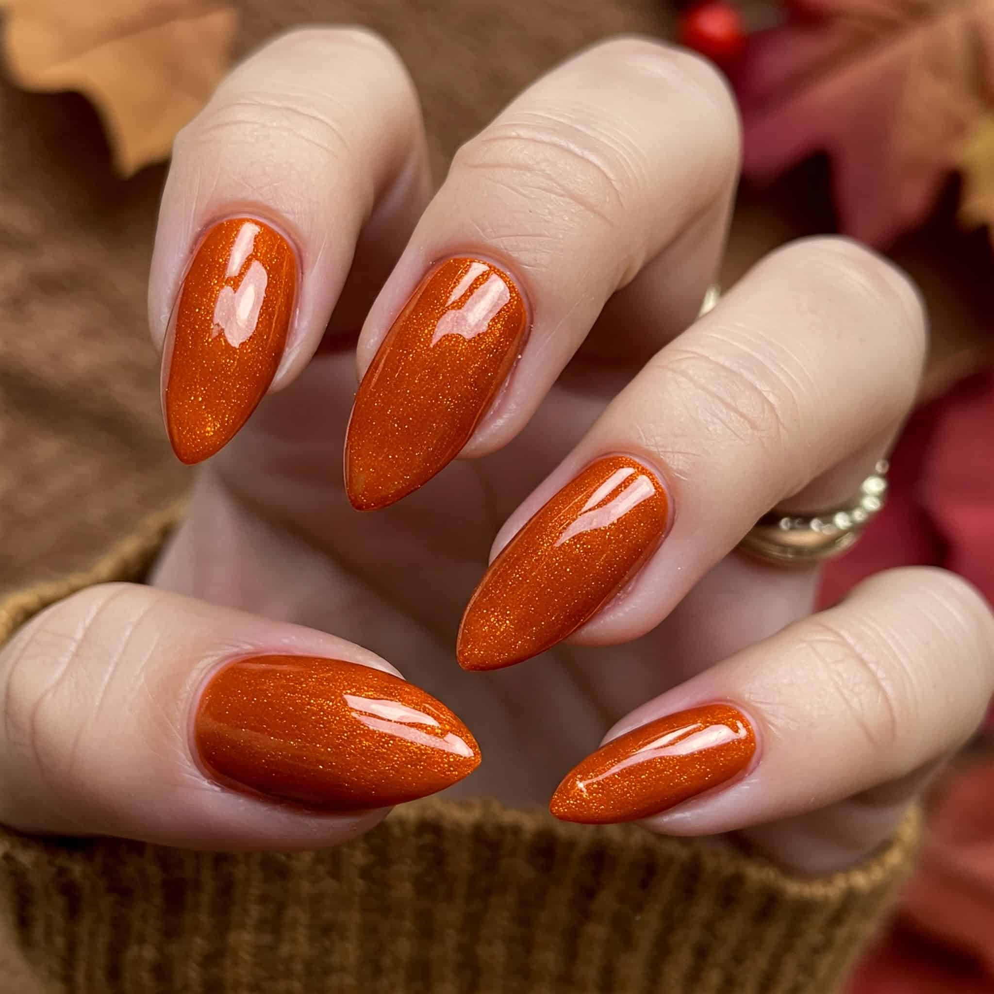 Close-up of a hand with glossy, pointed, orange nails. The background features blurred autumn leaves, conveying a warm, fall-themed aesthetic.