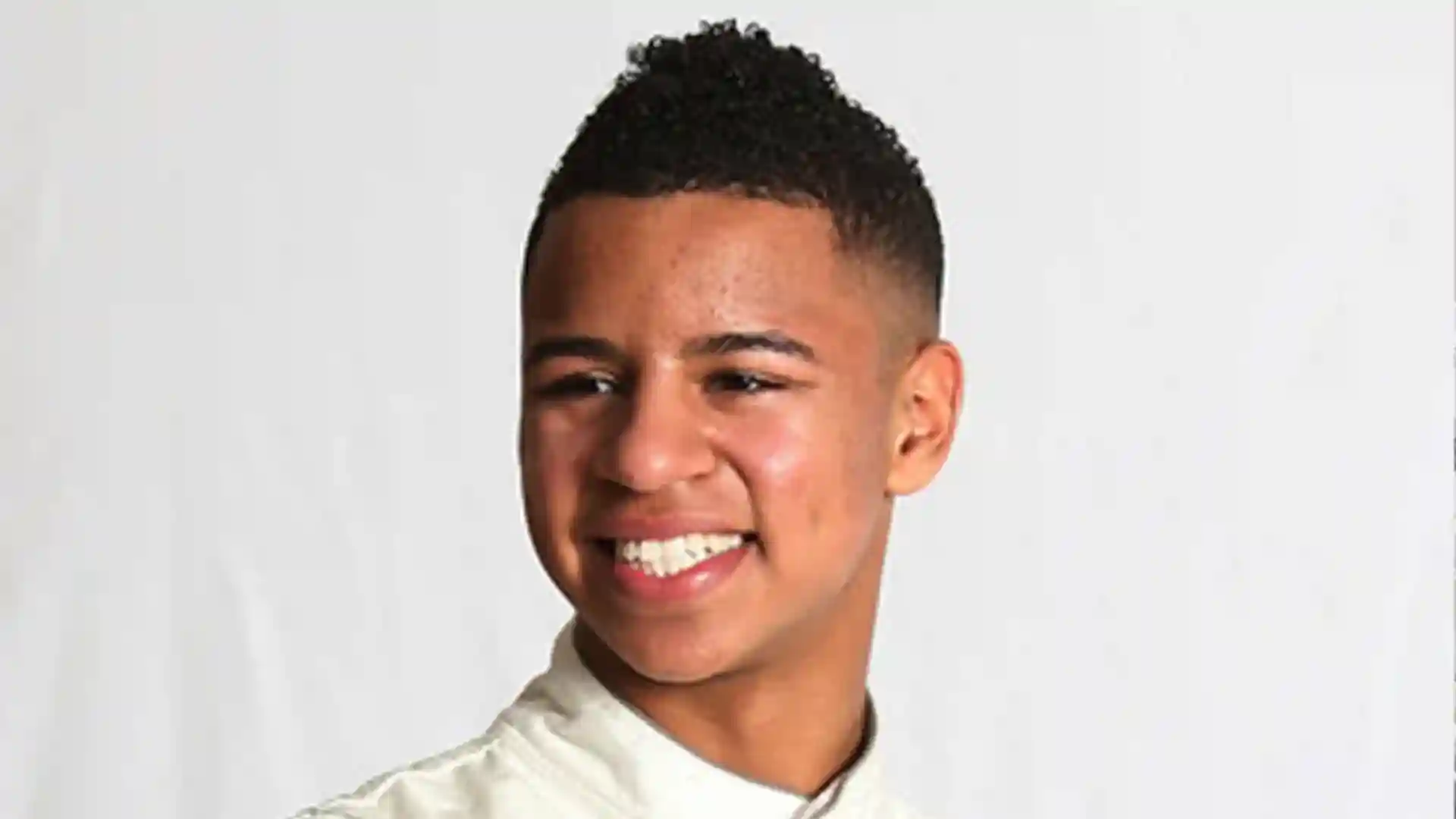 Smiling young man with a mohawk hairstyle wearing a light-colored collared shirt against a white background.