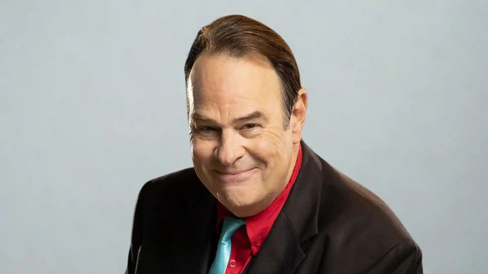 Portrait of Dan Aykroyd, actor and comedian, smiling in black suit, red shirt, and blue tie.
