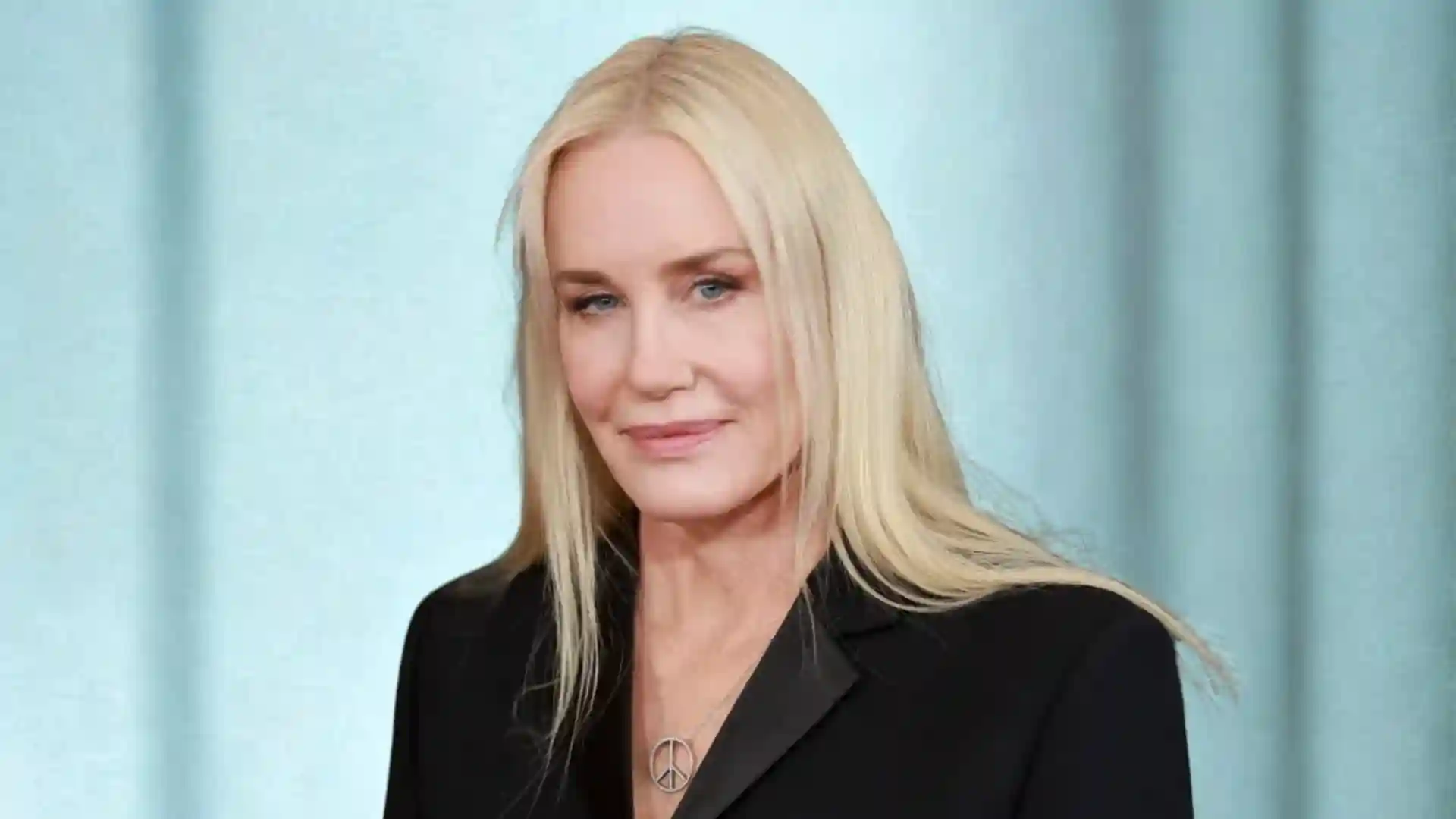 A blonde woman wearing a black blazer and a peace sign necklace smiles softly against a light blue backdrop.