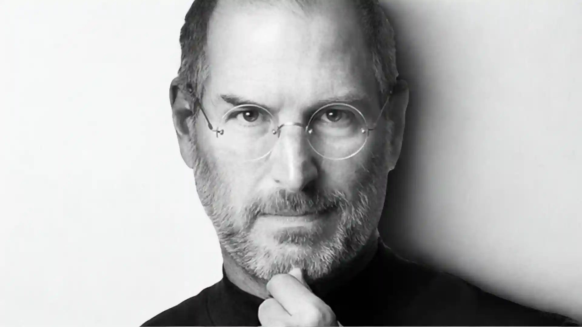 Portrait of Steve Jobs, in black turtleneck with round glasses and hand on chin.