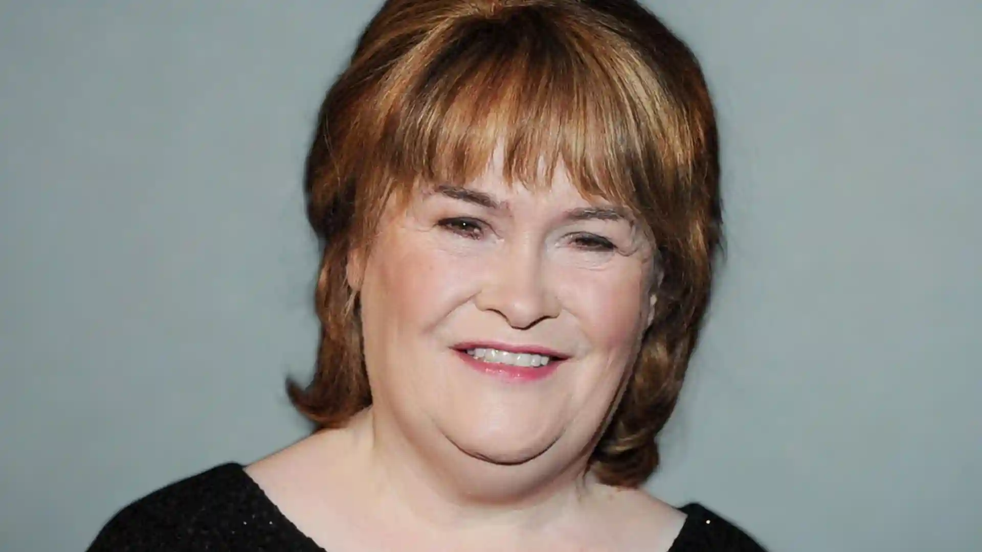 Close-up portrait of Susan Boyle smiling in front of a grey background.