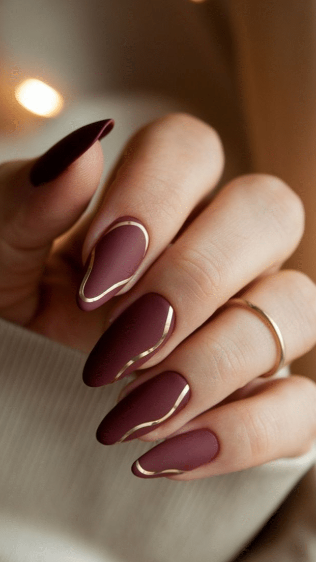 Almond-shaped nails in matte burgundy with gold swirl accents, creating a classy brown nail designs look.
