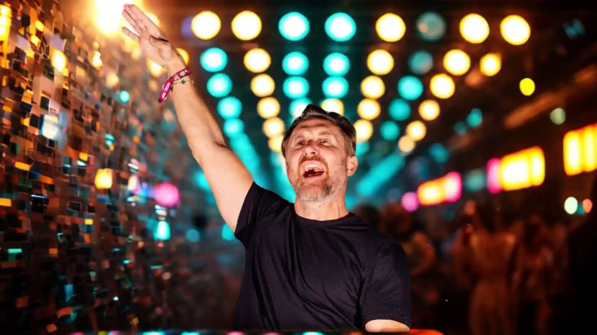 Dravis Guetta in black t-shirt with arm raised in front of disco ball wall and blurred colorful lights.