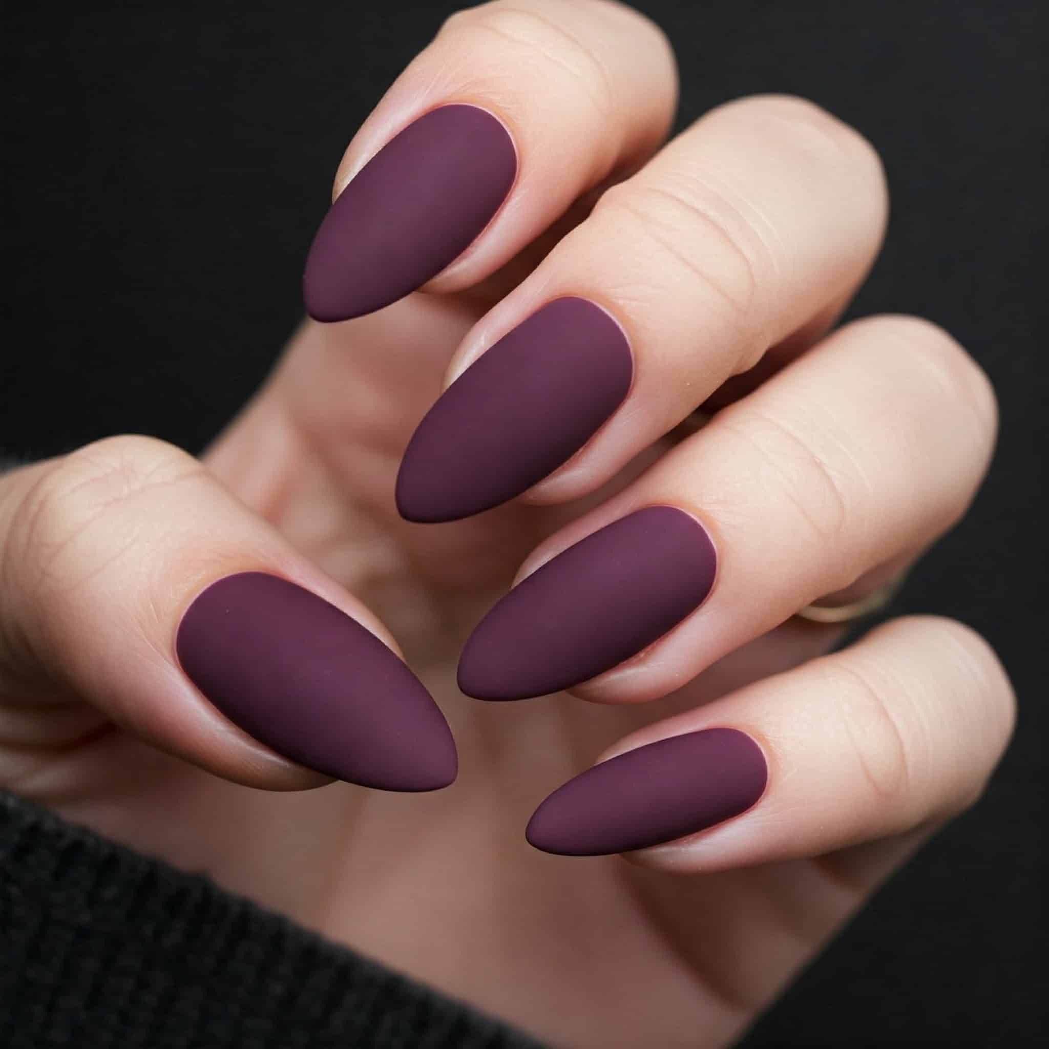 Close-up of a hand with matte plum-colored almond-shaped nails against a dark background.