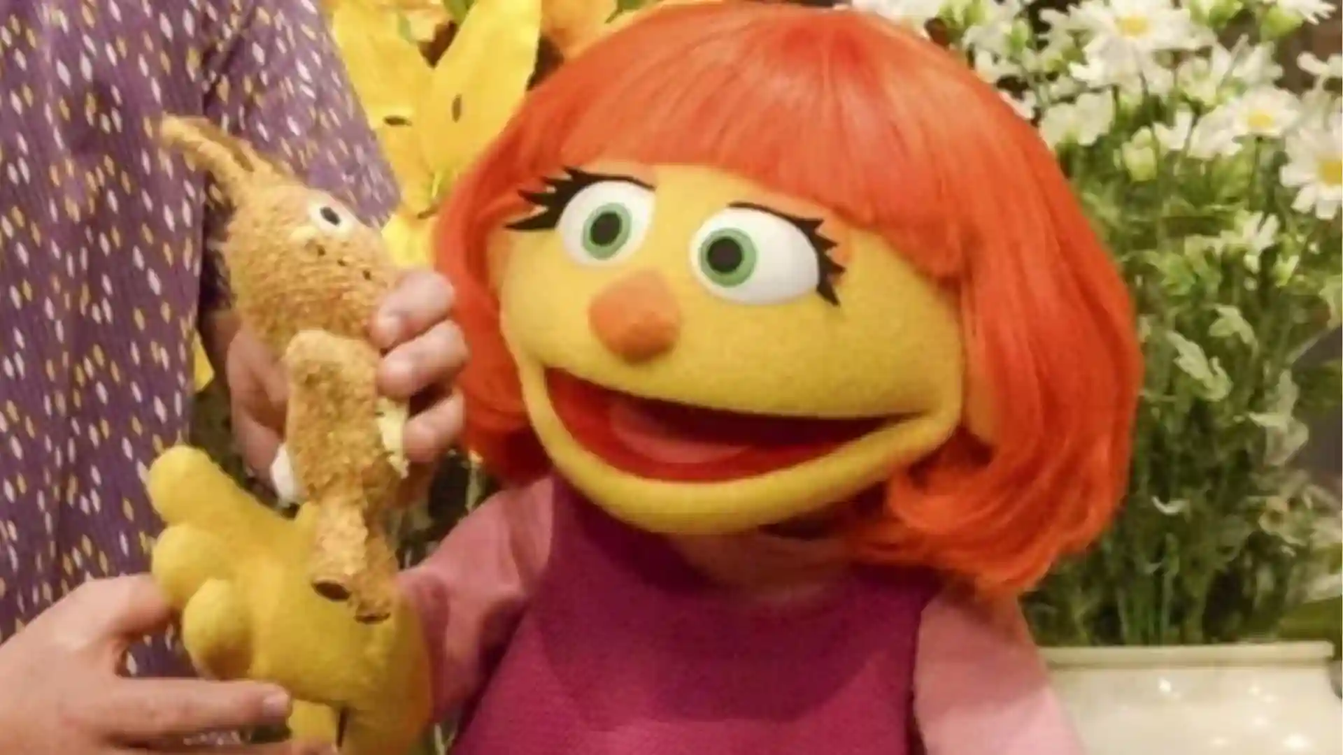 Red-haired puppet holding a stuffed rabbit, standing in front of flowers.