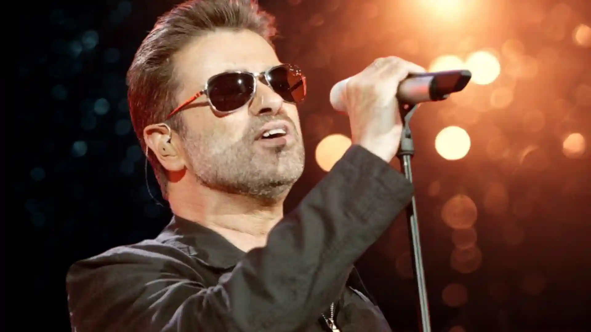 Singer George Michael performing on stage wearing sunglasses and holding a microphone.