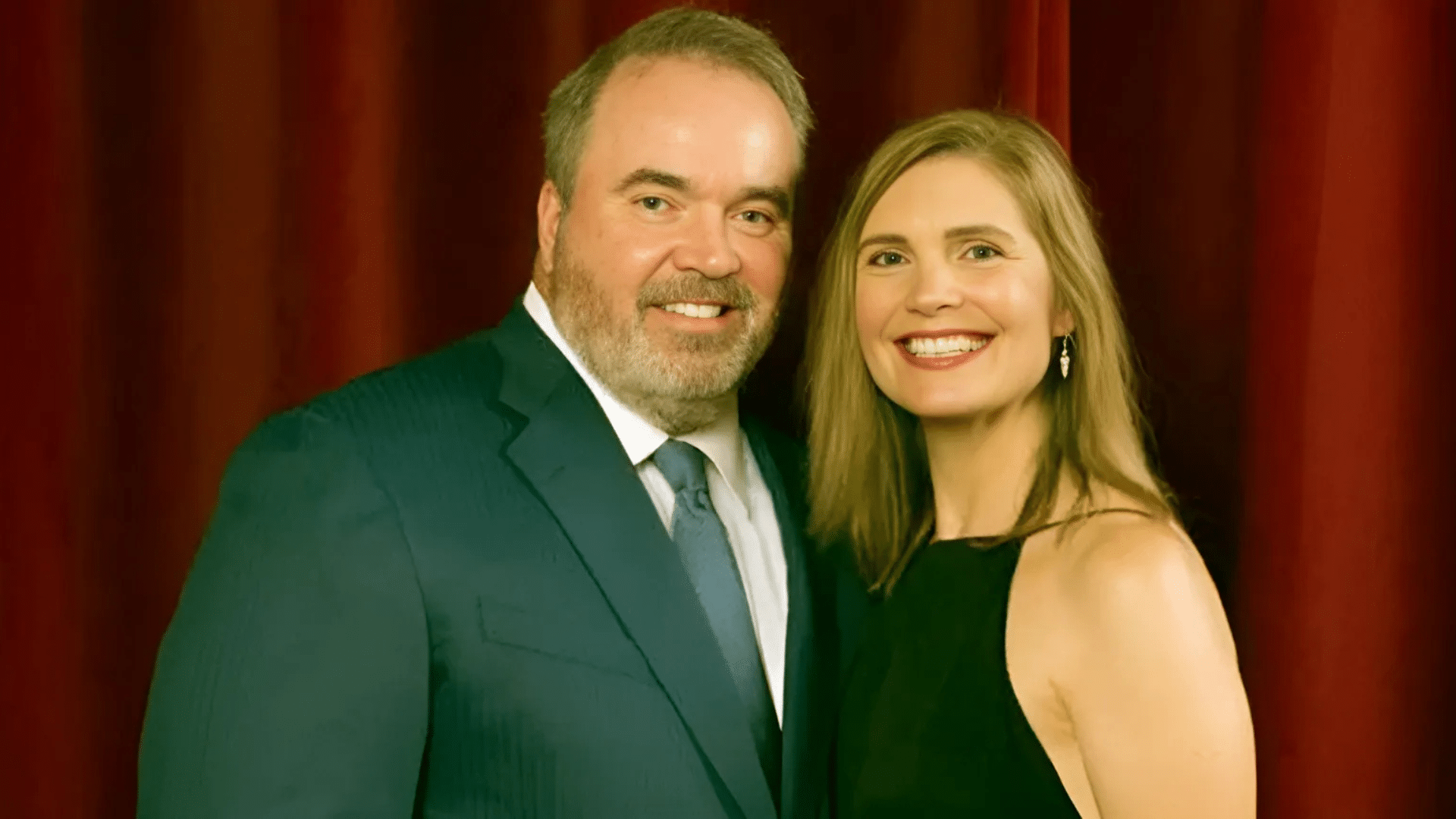 Jessica Kress posing with husband McCarthy in formal attire, both smiling in front of a red curtain backdrop.