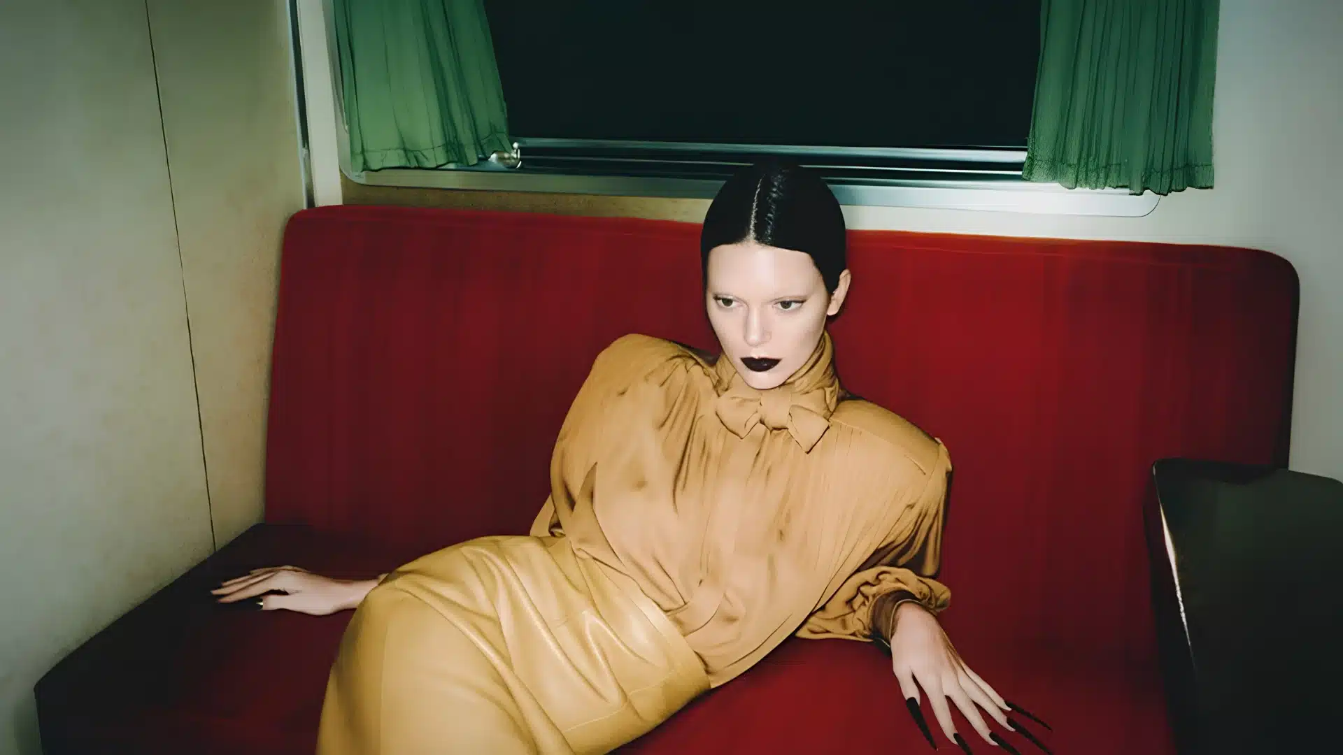 Model with dark lipstick and long black nails reclines on a red bench with Khaite's tan blouse and skirt