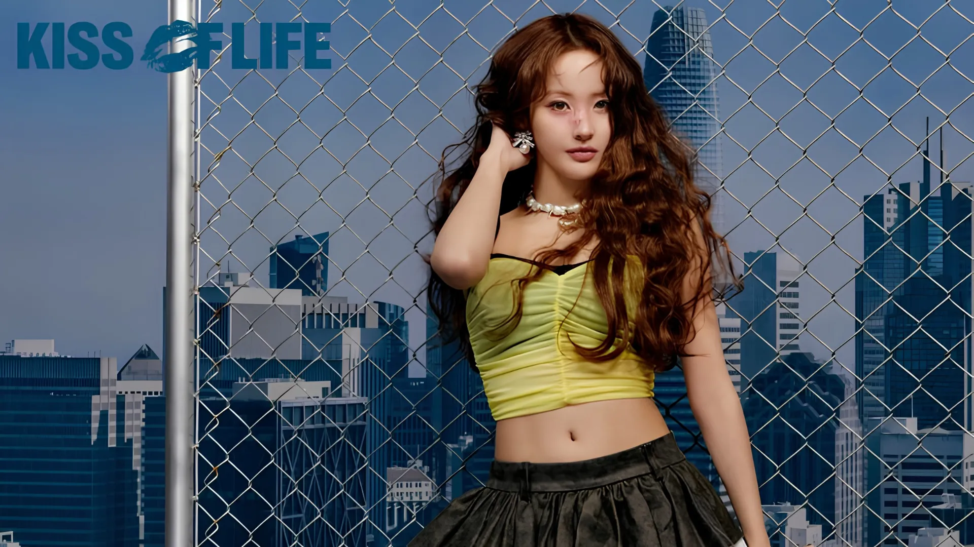 Kiss of Life member Belle with long, wavy brown hair poses in front of a fence with a cityscape in the background