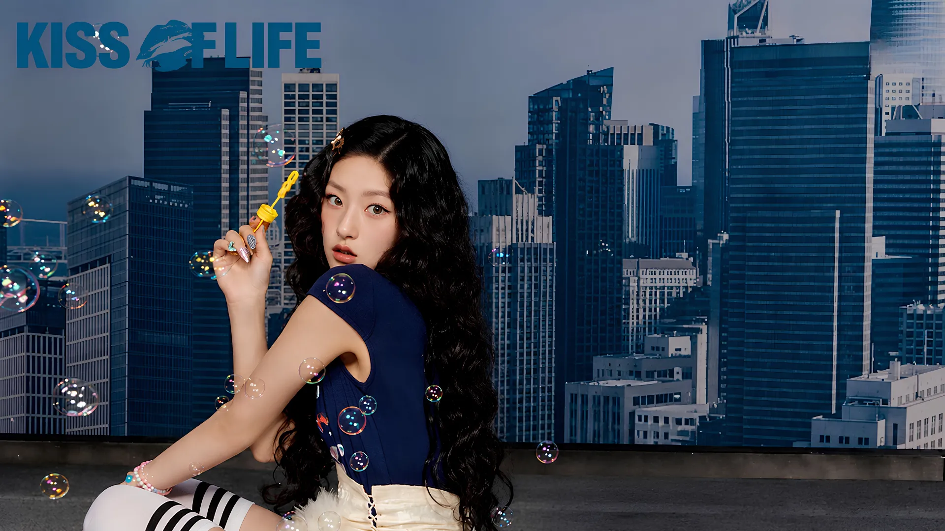 Kiss of Life member Hanuel long dark wavy hair blows bubbles in front of a cityscape