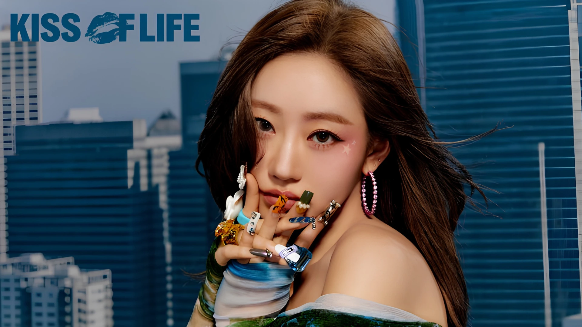 Kiss of Life member Julie with elaborate fingernails and pink hoop earrings, posing in front of a blurred cityscape