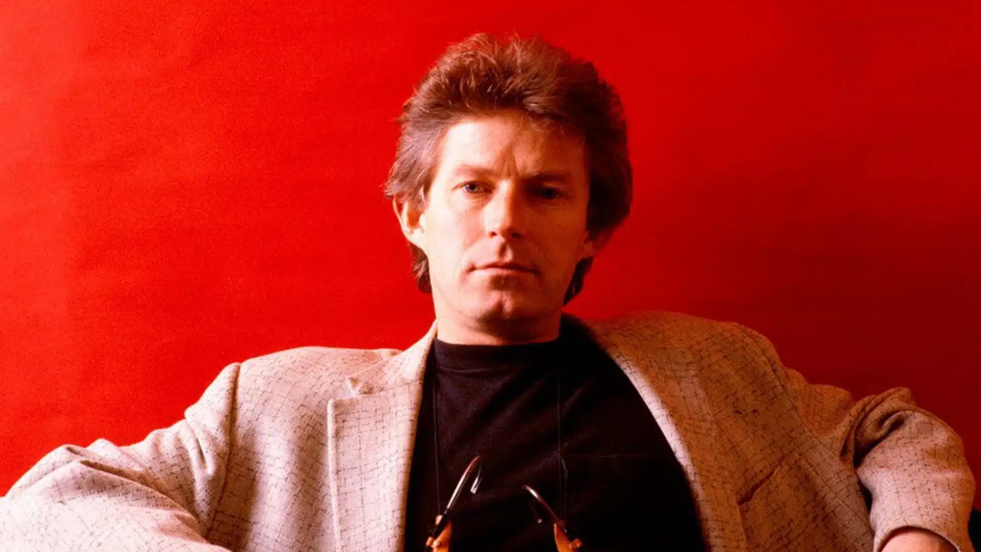 Don Henley with light brown hair wearing a blazer and glasses against a red background