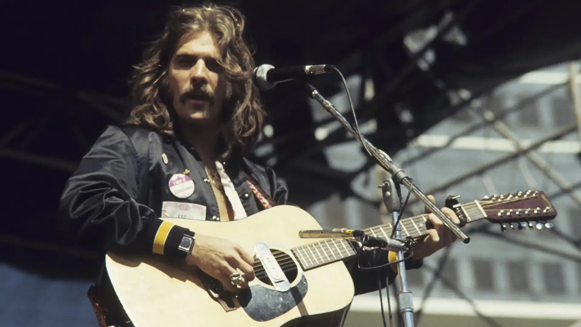 Glenn Frey plays a 12 string acoustic guitar on stage, singing into a microphone