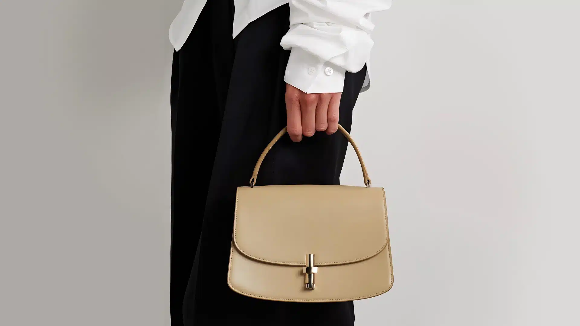 A person in a white shirt and black pants holds a smooth, light brown leather handbag with a gold clasp