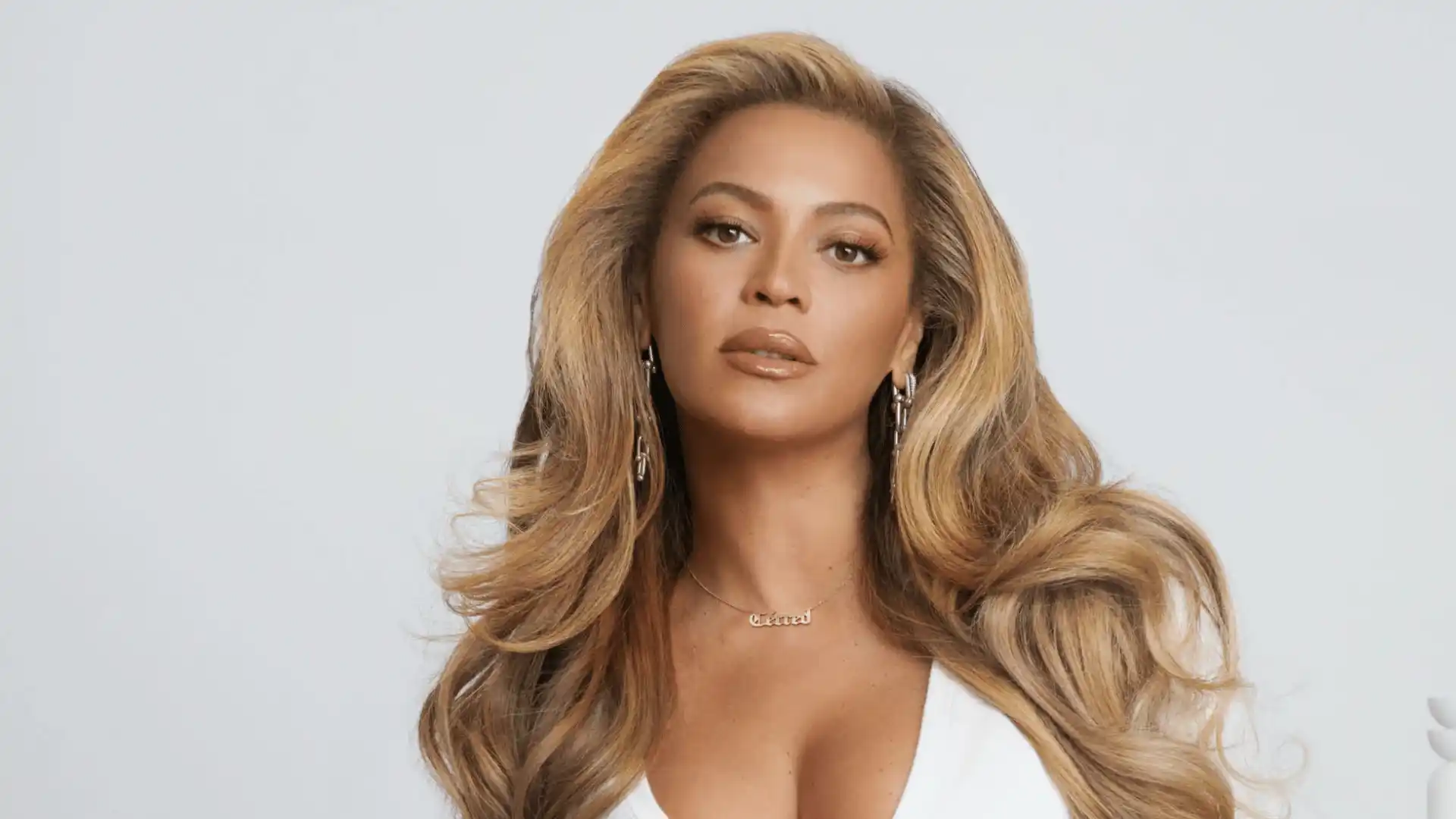 Beyonce with long blonde hair, wearing a white outfit and earrings, looking directly at the camera against a light background