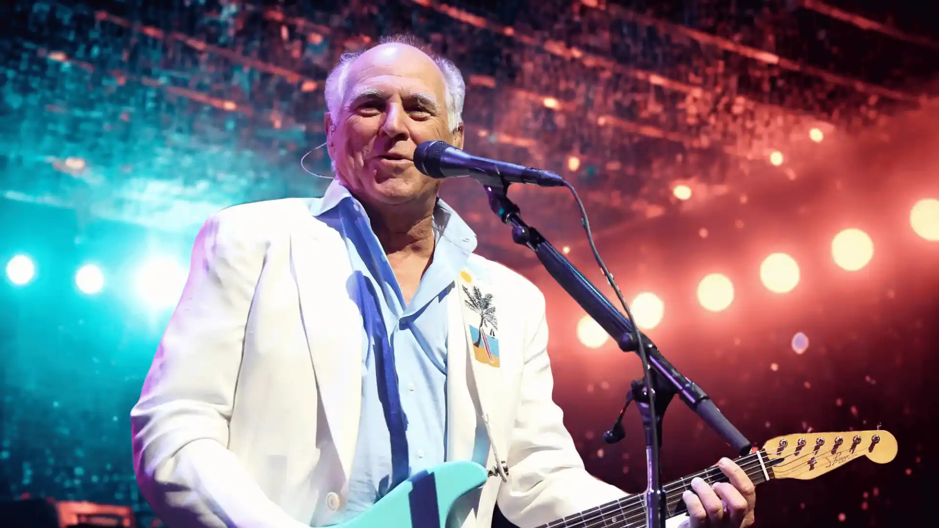 Jimmy Buffett performing on stage singing into a microphone and playing guitar under lights.