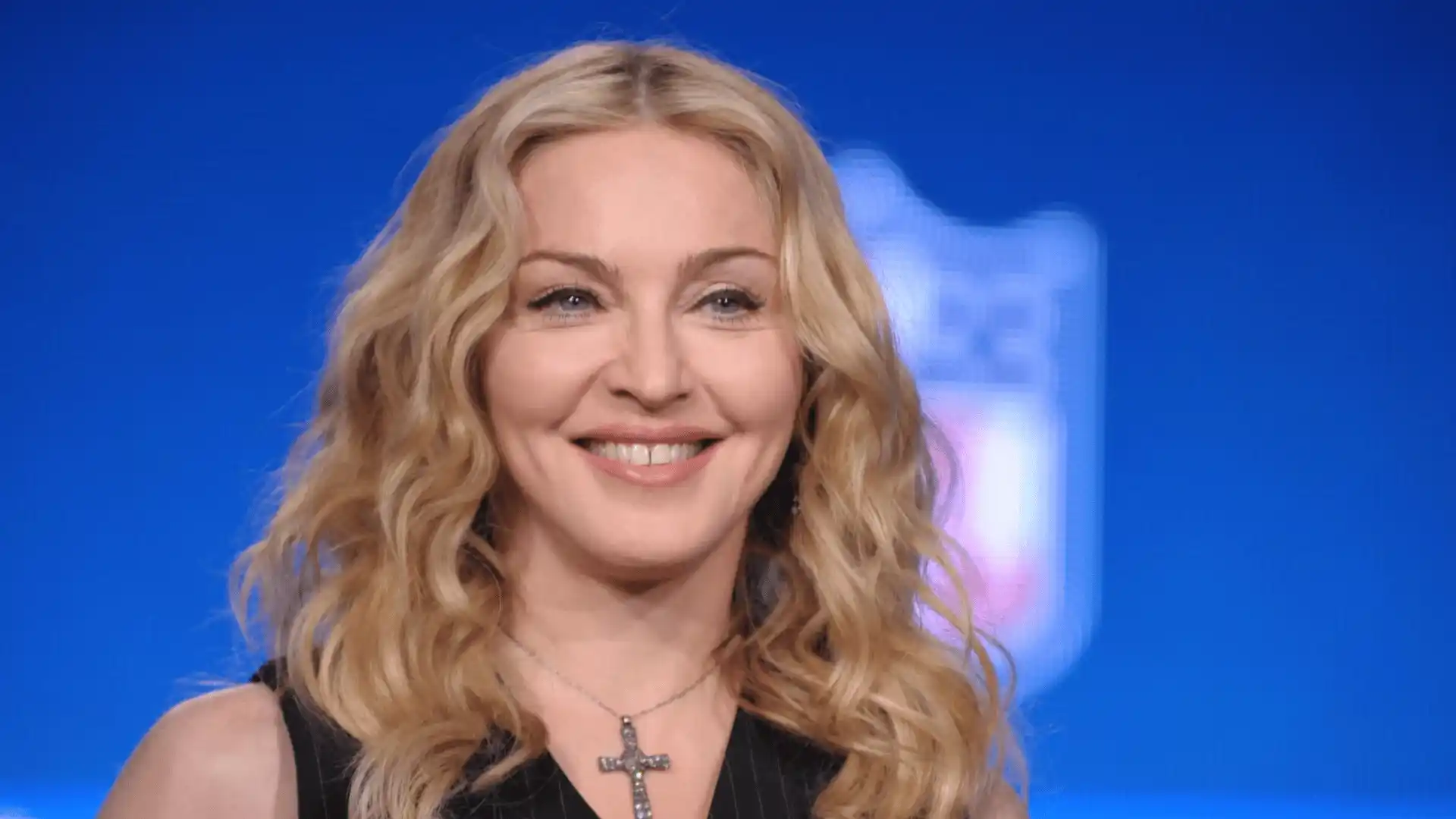 Madonna smiling with wavy blonde hair and cross necklace against a blue studio background portrait
