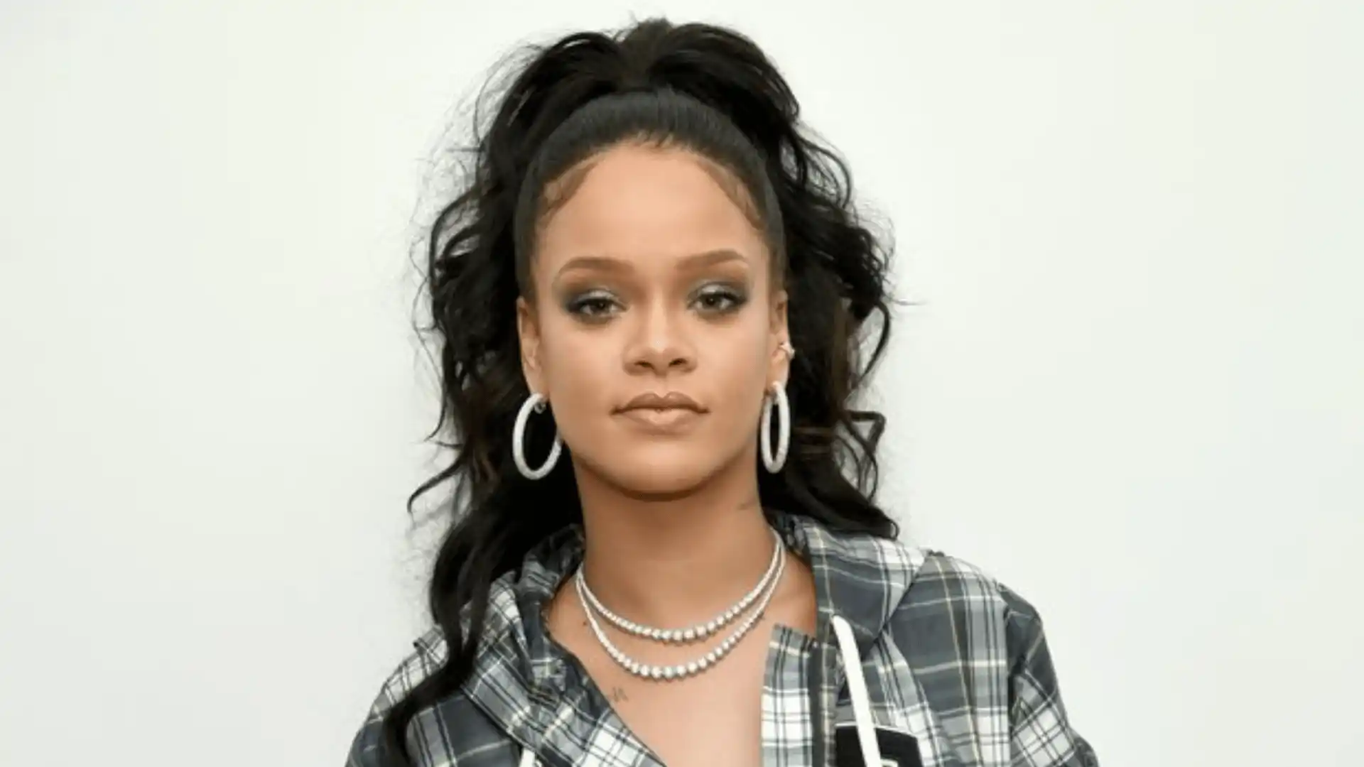 Rihanna with dark hair in a high ponytail, hoop earrings, layered necklaces, and a plaid shirt against a plain background