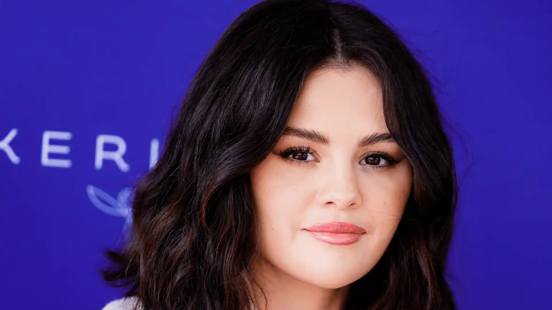 Selena Gomez poses against a blue background with faint text.