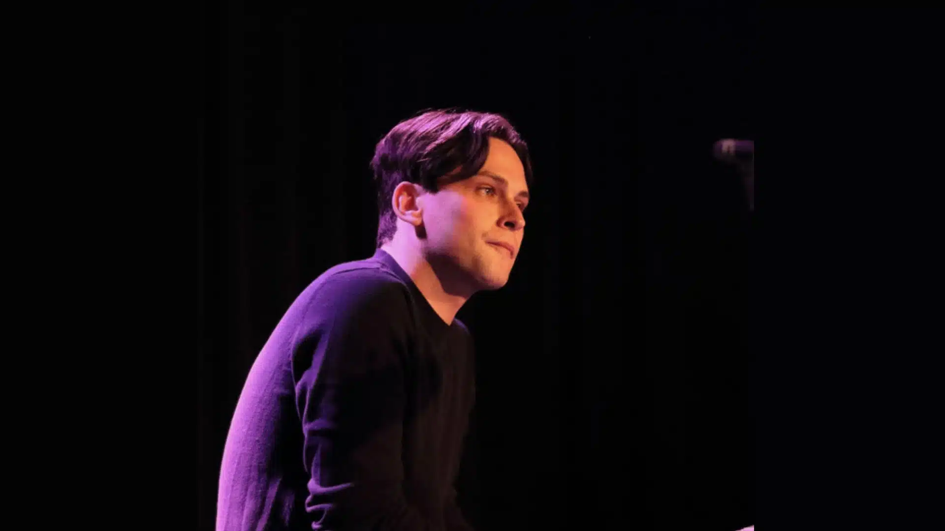 Edward James wearing a black shirt is illuminated by purple stage lighting against a black backdrop