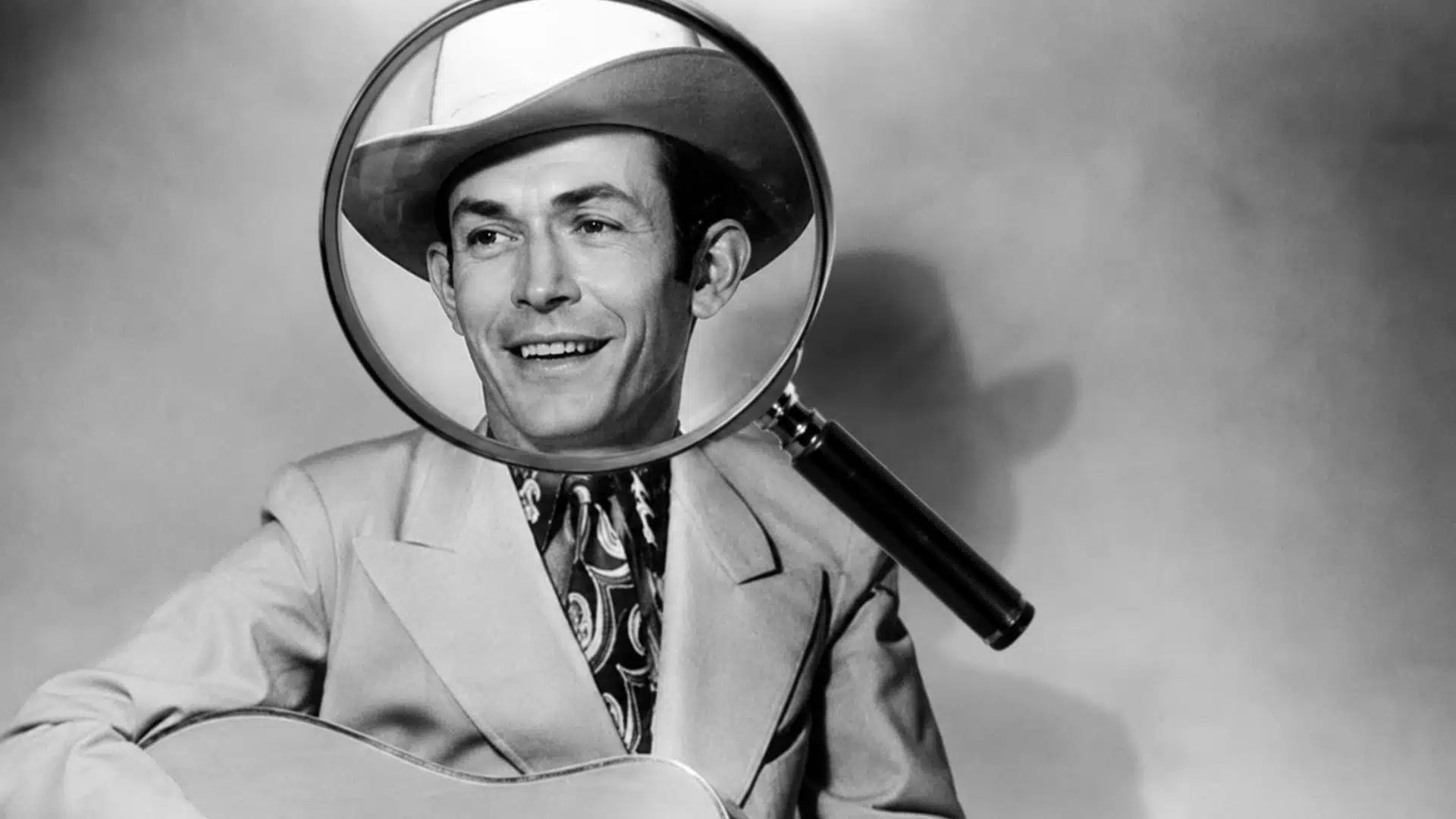 Black and white photo of Hank Williams in a cowboy hat and suit holding a guitar, with a magnifying glass over his face